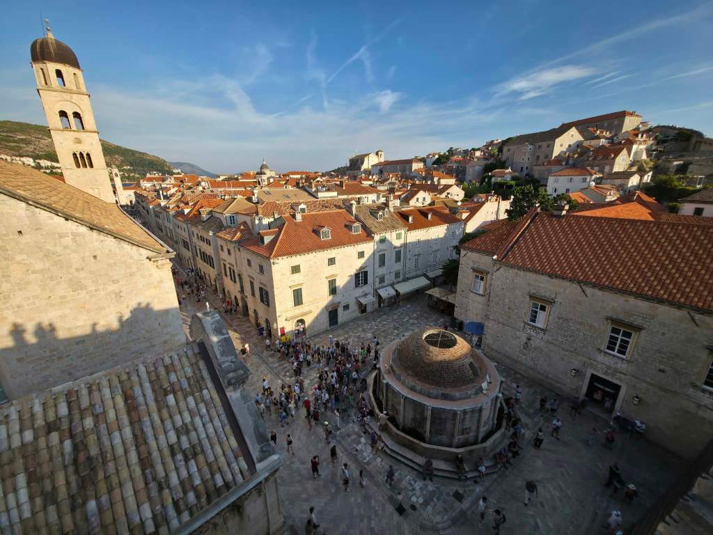 A panoramic view of a historic town square in Dubrovnik, Croatia, showcasing terracotta-roofed buildings, a central fountain, and a crowd of people enjoying the area.