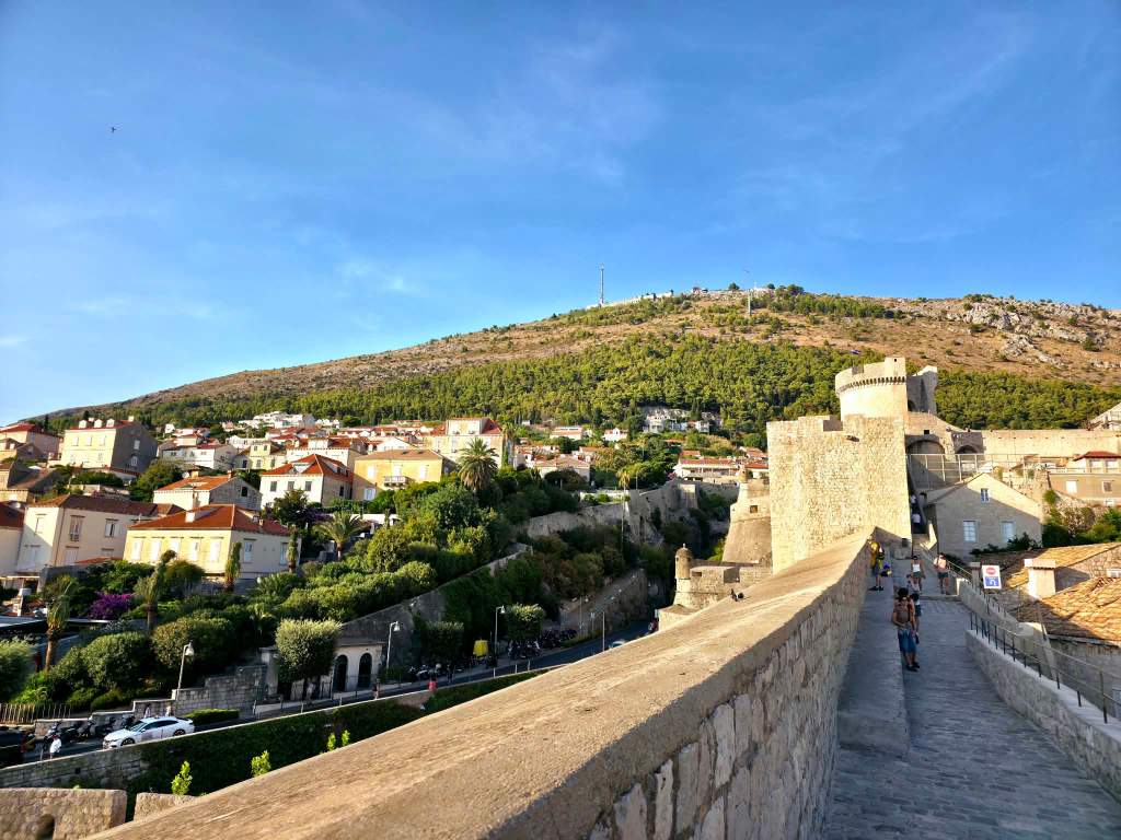 A scenic view of a hillside town with homes, greenery, and a historic stone wall, under a clear blue sky.