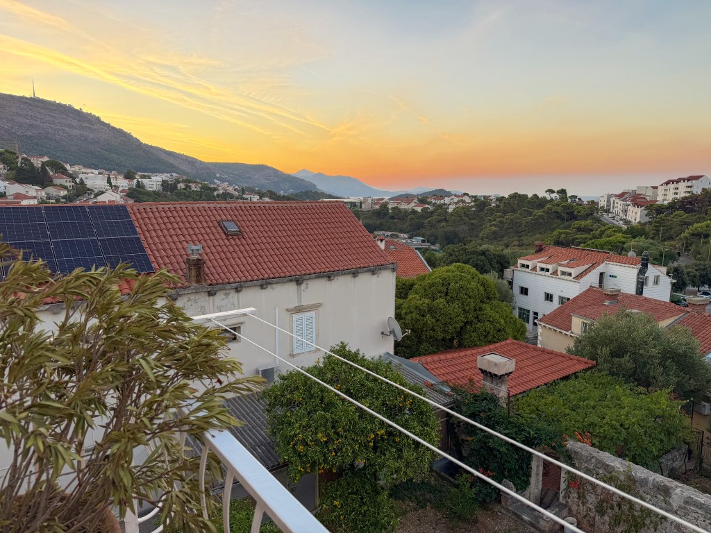 A scenic view of rooftops with red tiles, solar panels, and lush greenery, overlooking a mountainous landscape at sunset with vibrant orange and yellow hues in the sky.