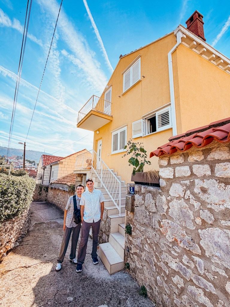 Two young men standing outside a yellow house with white shutters and a balcony, on a sunny day with a blue sky and clouds.