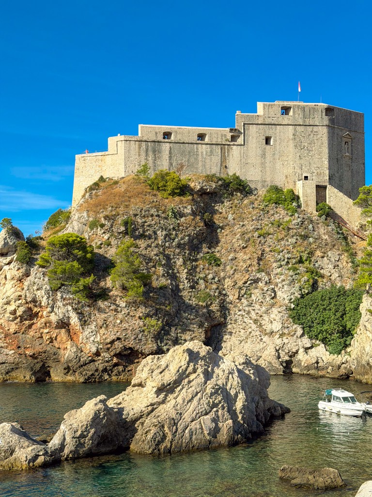 A historic stone fortress perched on a rocky cliff overlooking a calm blue sea, with a small boat anchored nearby and clear blue skies above.