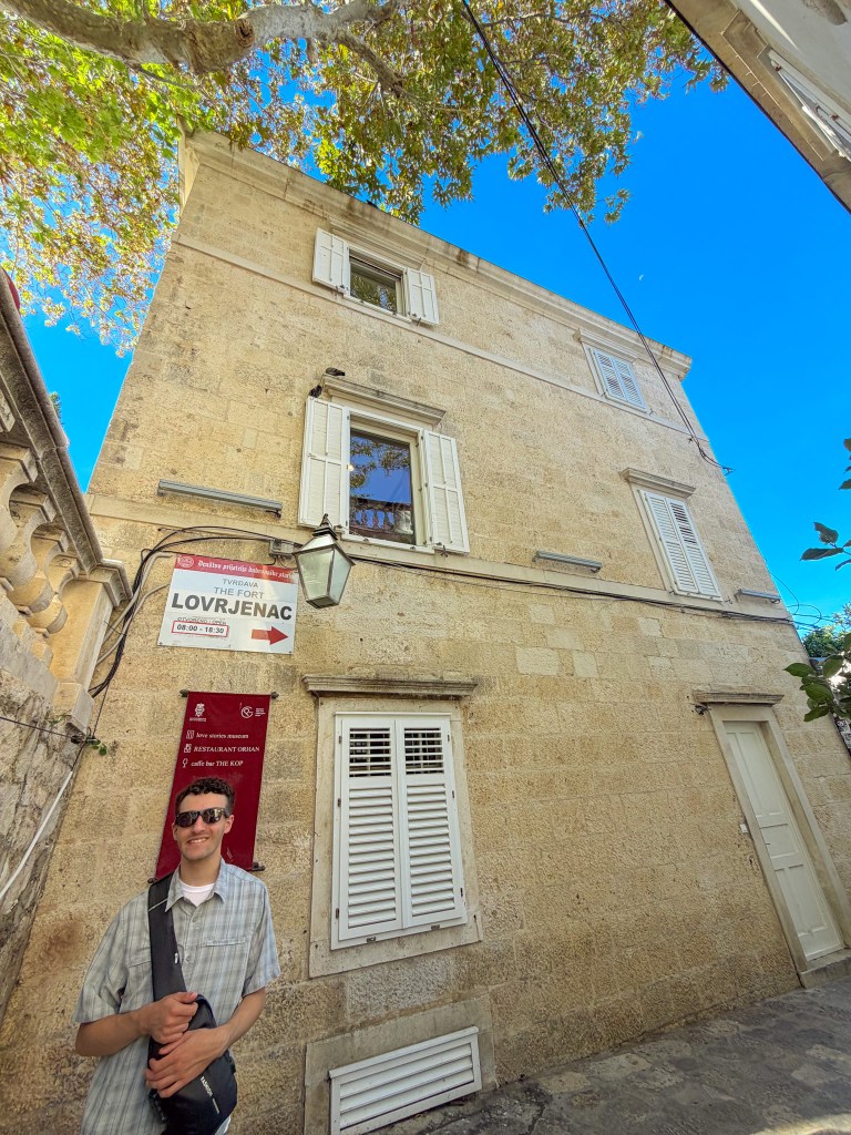 A man with sunglasses stands in front of a light-colored stone building with white shutters, surrounded by greenery and a clear blue sky. A sign indicates the direction to Fort Lovrijenac.