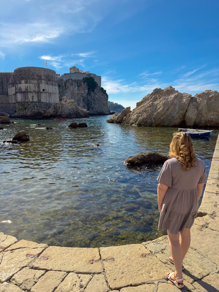 A person standing by the water's edge, overlooking a rocky coastline with historical buildings in the background under a clear blue sky.