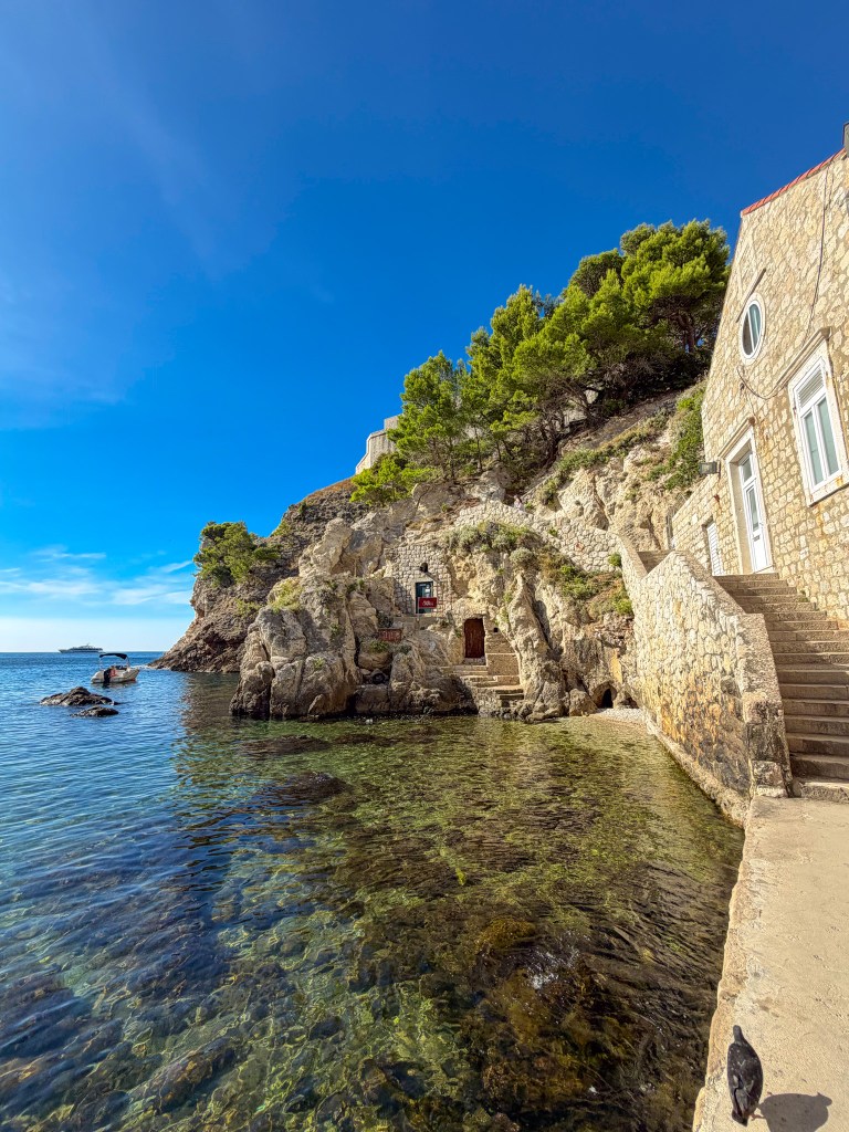 Scenic coastal view featuring a rocky shore with clear water, a stone building, and greenery on a hillside under a blue sky.