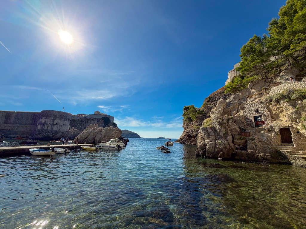 A scenic view of a calm bay with small boats moored along a rocky shoreline, framed by cliffs and a clear blue sky. The sun shines brightly overhead.
