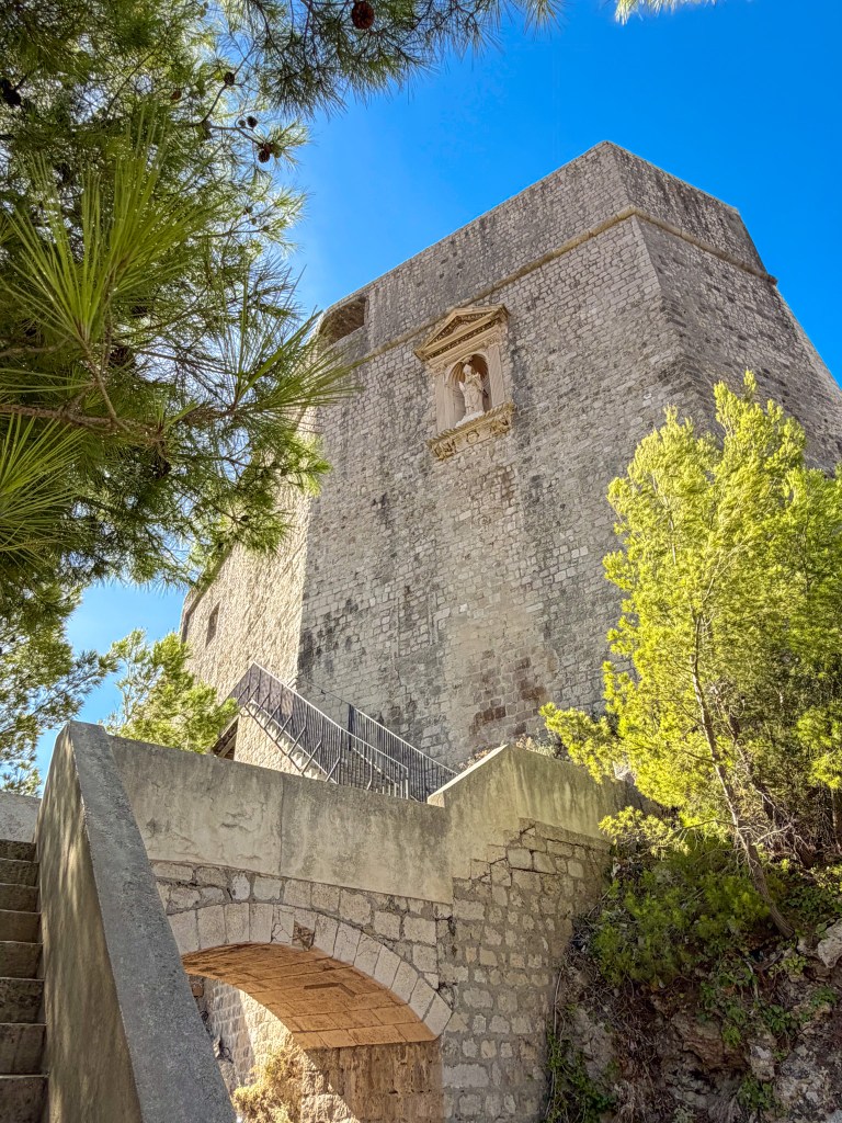 A stone fortress viewed from below, with an ornate facade featuring a statue and surrounded by lush greenery and blue skies.
