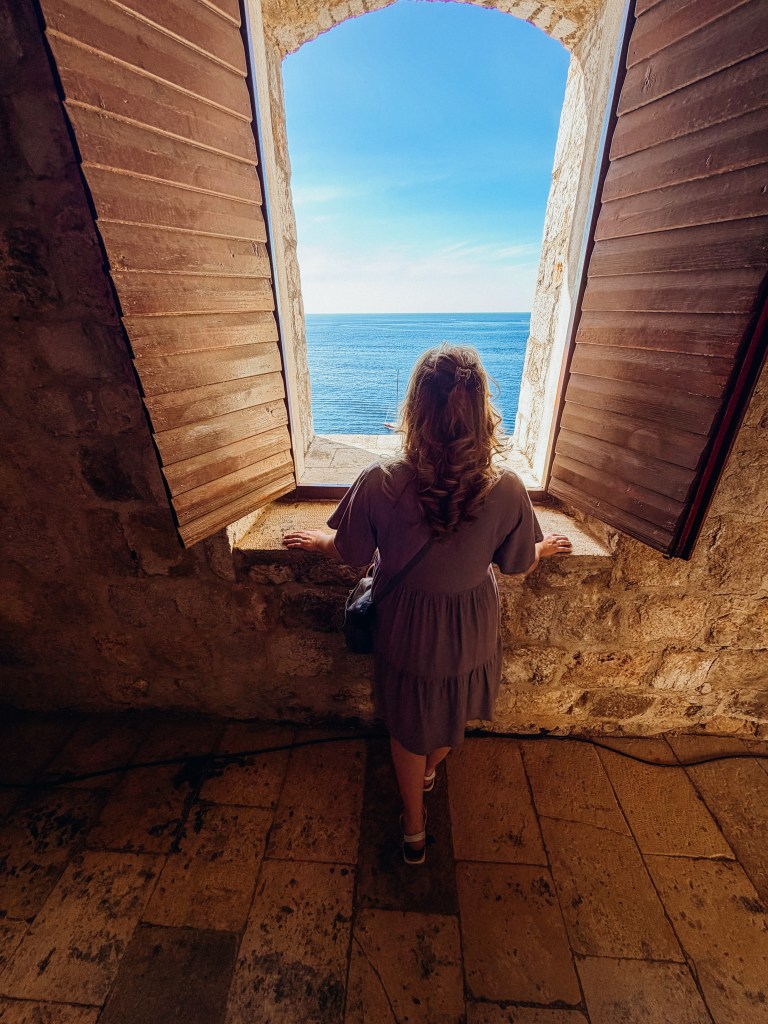 A person stands at a window with wooden shutters open, overlooking the ocean and a clear blue sky.