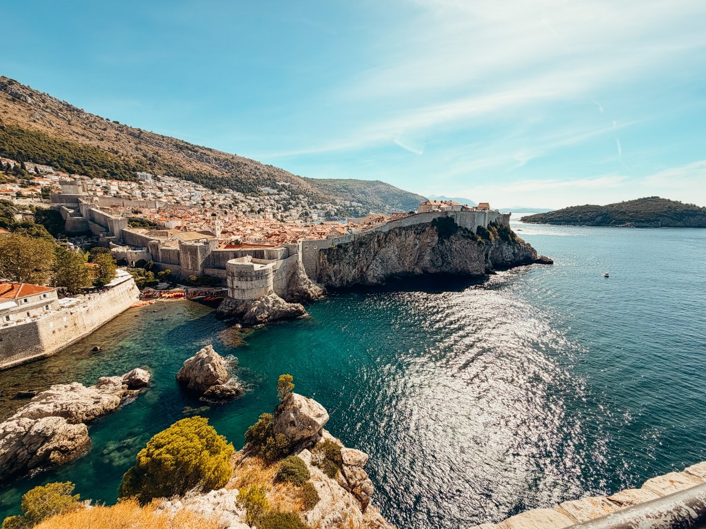 A panoramic view of Dubrovnik, Croatia, featuring ancient city walls along the coastline and the blue waters of the Adriatic Sea, with green hills in the background.