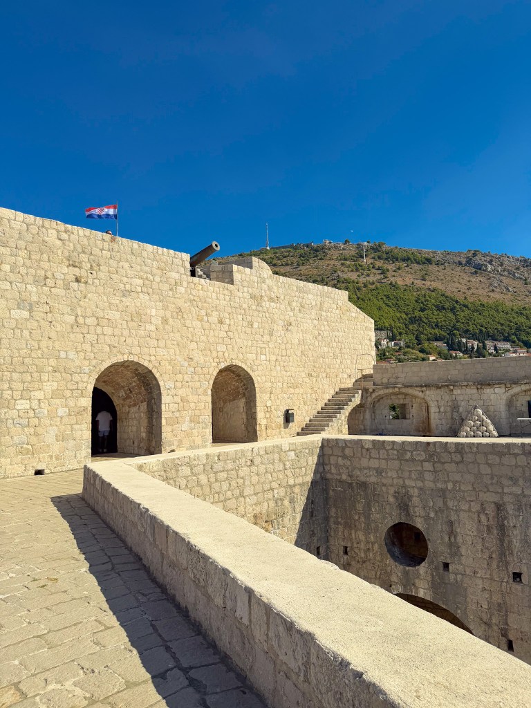 View of a stone fortress with arches and a Croatian flag, overlooking a hillside under a clear blue sky.