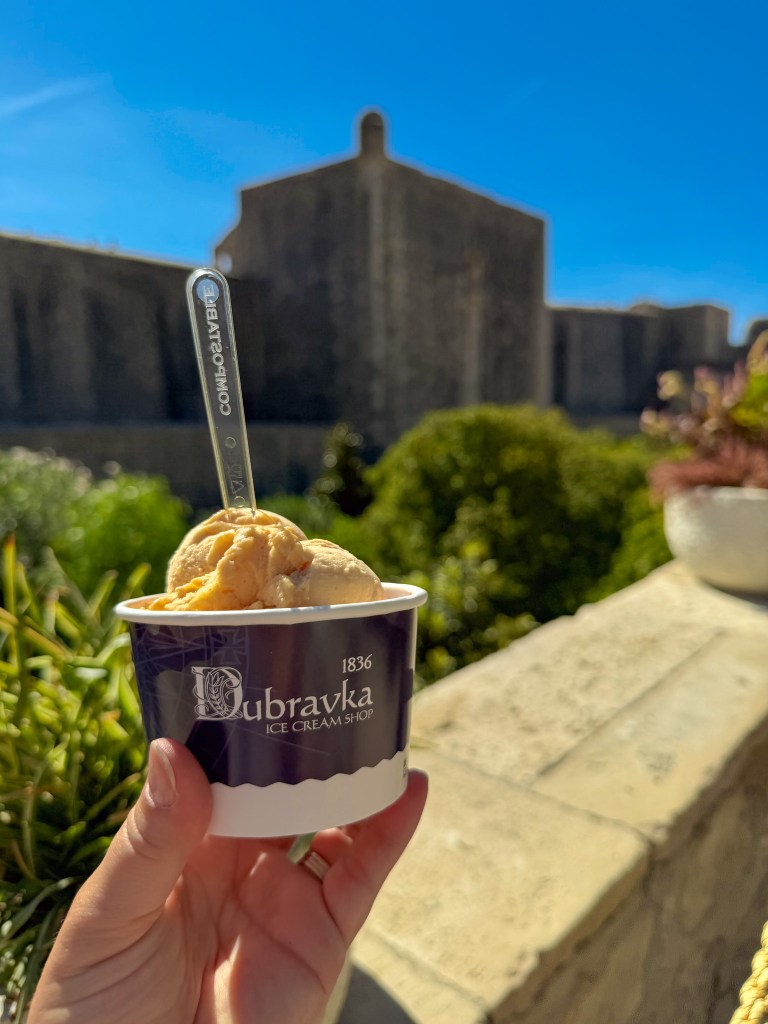 A hand holding a cup of ice cream from Dubravka Ice Cream Shop with a scenic background featuring a stone building and clear blue sky.