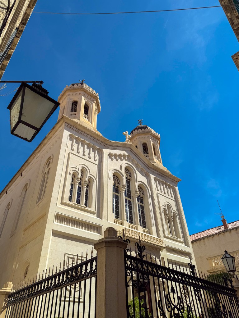 A large, white church with intricate architectural details, featuring two towers and decorative windows, set against a clear blue sky.