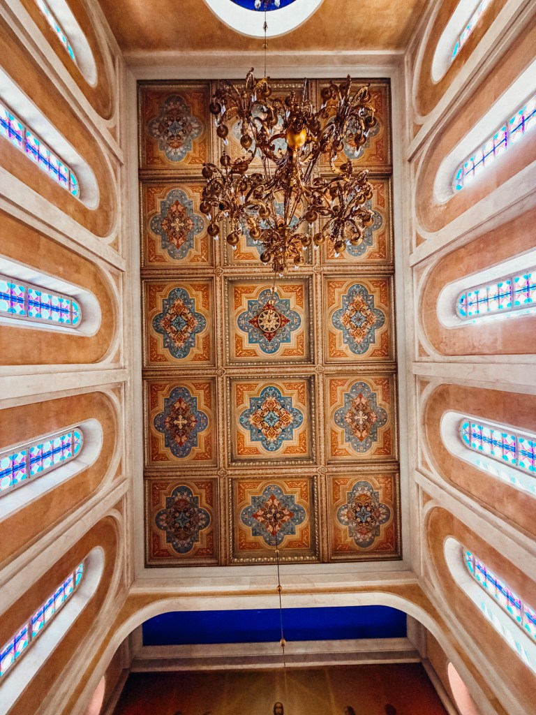 Interior view of a church ceiling featuring ornate patterned tiles, a decorative chandelier, and colorful stained glass windows.