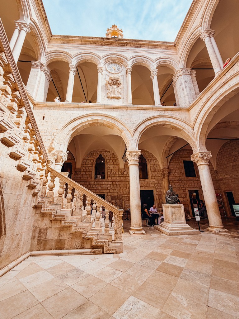 Interior view of a historic building featuring a grand staircase, arched columns, and a statue in the center. Natural light illuminates the space from above.