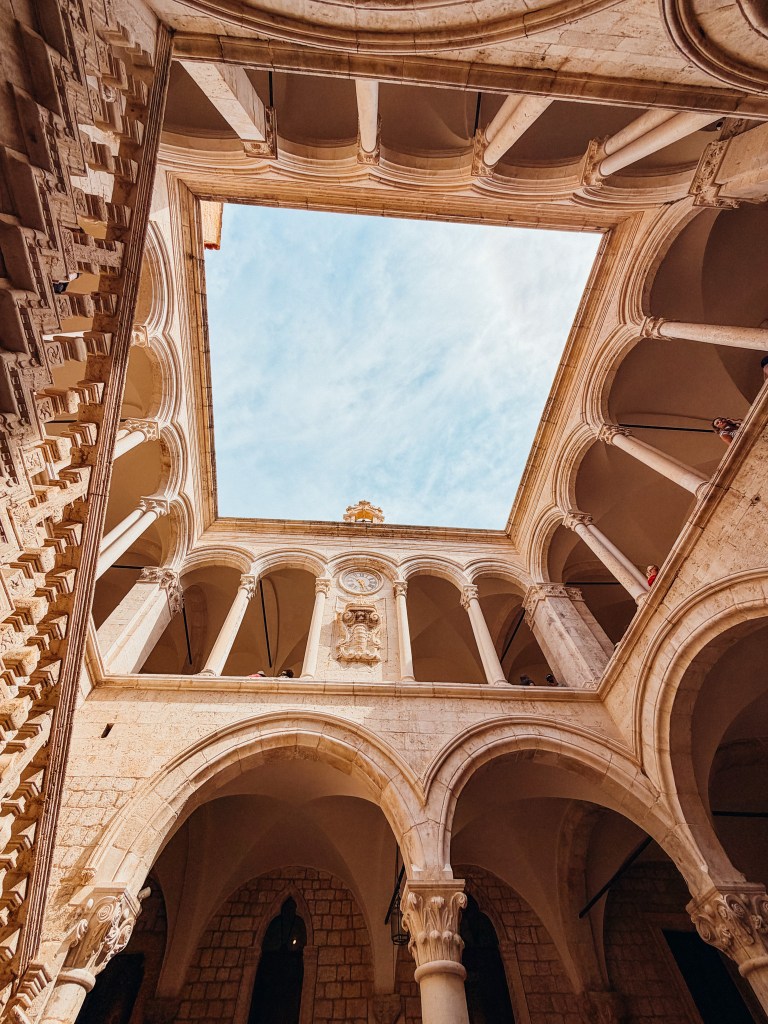View of a stone courtyard with arched columns and a blue sky above, showcasing a historic architectural design.