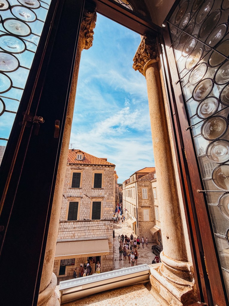 View from a window with decorative glass, showcasing a stone street bustling with people and historic buildings under a blue sky.