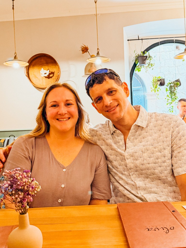 A smiling couple sitting at a wooden table in a cozy restaurant, with a small vase of flowers in the foreground and a menu in front of them.