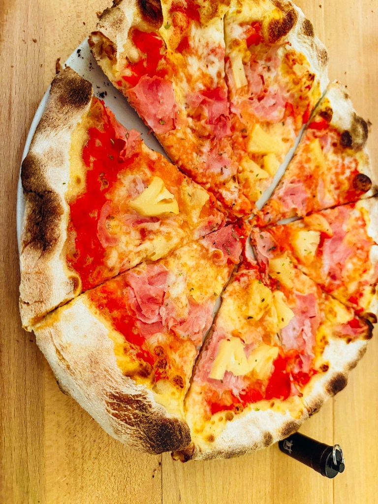 A freshly baked pizza topped with ham and pineapple, cut into slices, placed on a wooden surface.
