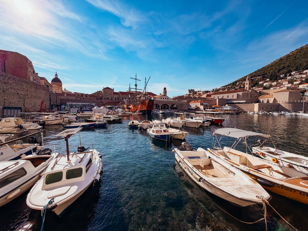 Harbor scene with various boats anchored in calm waters, surrounded by historic buildings and hills under a clear blue sky.
