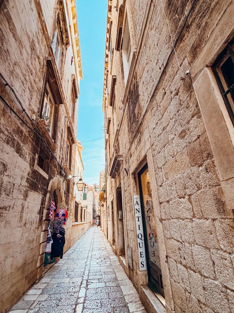 Narrow cobblestone alleyway between historic buildings with a shop displaying antiques on the right and a person browsing on the left under a colorful display.