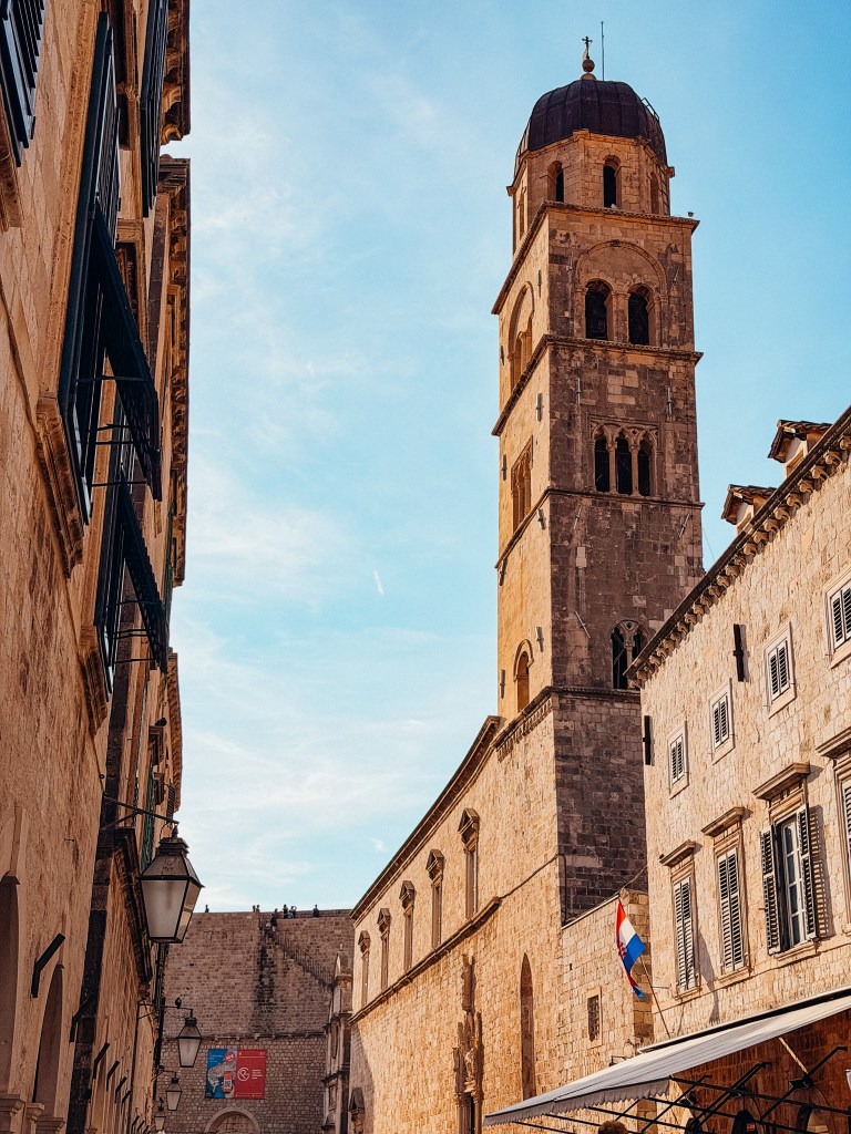 narrow street lined with historic stone buildings, featuring a tall bell tower against a blue sky